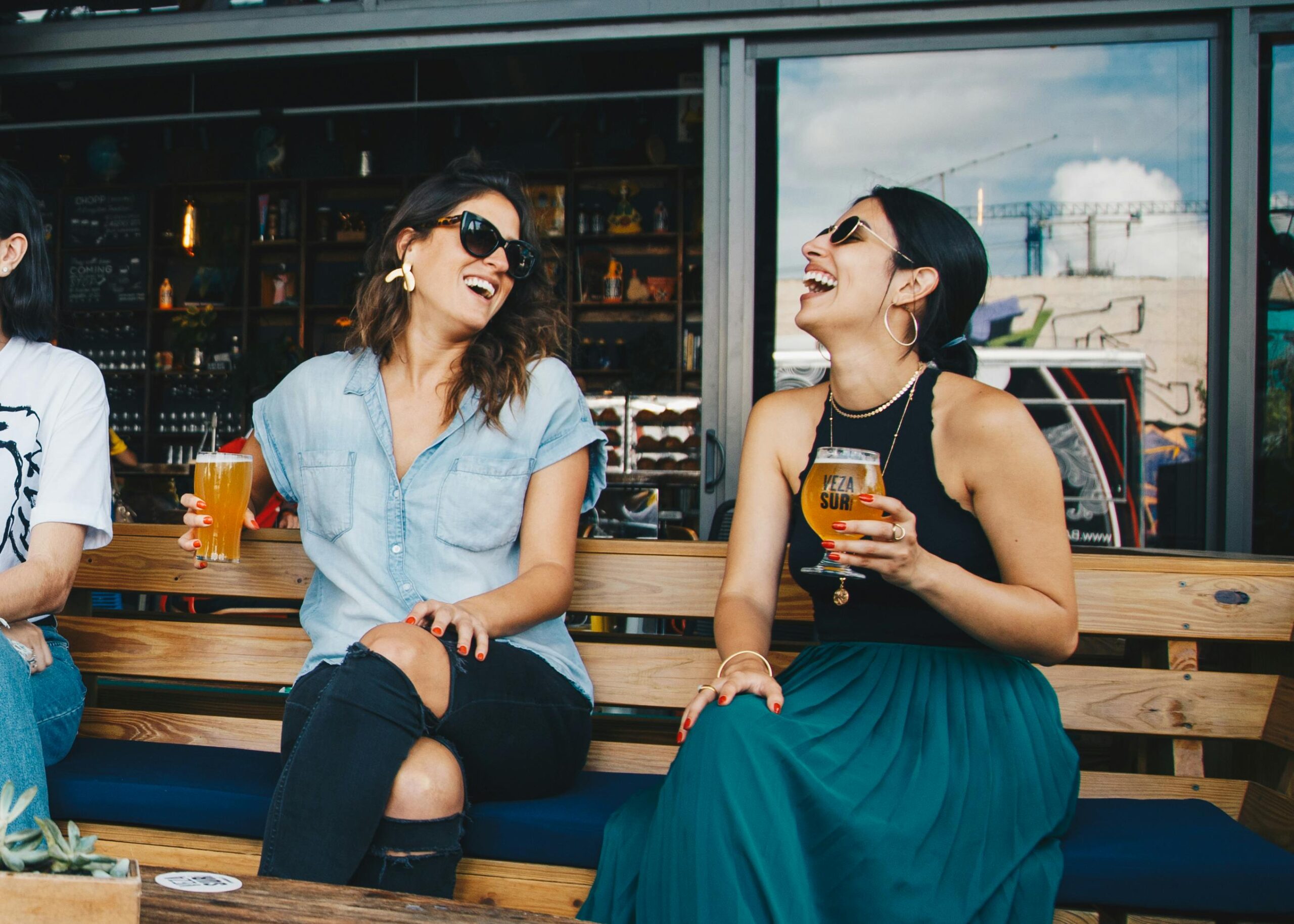 Fonctionnalités entreprises Two women laughing and enjoying drinks outdoors at a trendy bar, creating a vibrant and social atmosphere.